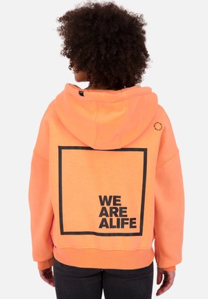 Personne aux cheveux bouclés portant un sweat à capuche orange avec un grand carré noir et un texte en gras "WE ARE ALIFE" au dos.