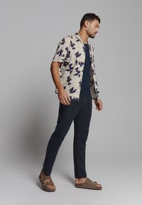 Korte mouwen crèmekleurig shirt met blauw bloemenpatroon, marineblauwe t-shirt, donkere broeken en bruine sandalen. Het materiaal lijkt lichtgewicht en casual te zijn.