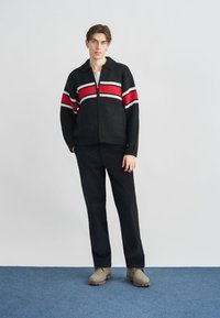Black knitted cardigan with red and white stripes, featuring a zip closure and a collar. Paired with black pants and beige boots.