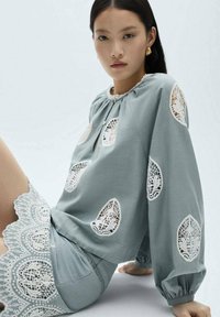 Blusa azzurro chiaro con dettagli in pizzo e finitura testurizzata, caratterizzata da un collo rotondo e maniche lunghe. Dettagli in pizzo sulle maniche e sull'orlo.