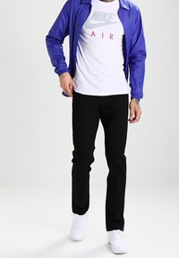 Modellen har på sig en blå lättviktsjacka över en vit Nike T-shirt med grå logotyp, svarta byxor och vita sneakers.