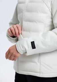 Biała puchowa kurtka z zamkiem błyskawicznym, posiadająca czarną naszywkę z logo na mankiecie oraz miękką wewnętrzną tkaninę na nadgarstku dla komfortu.