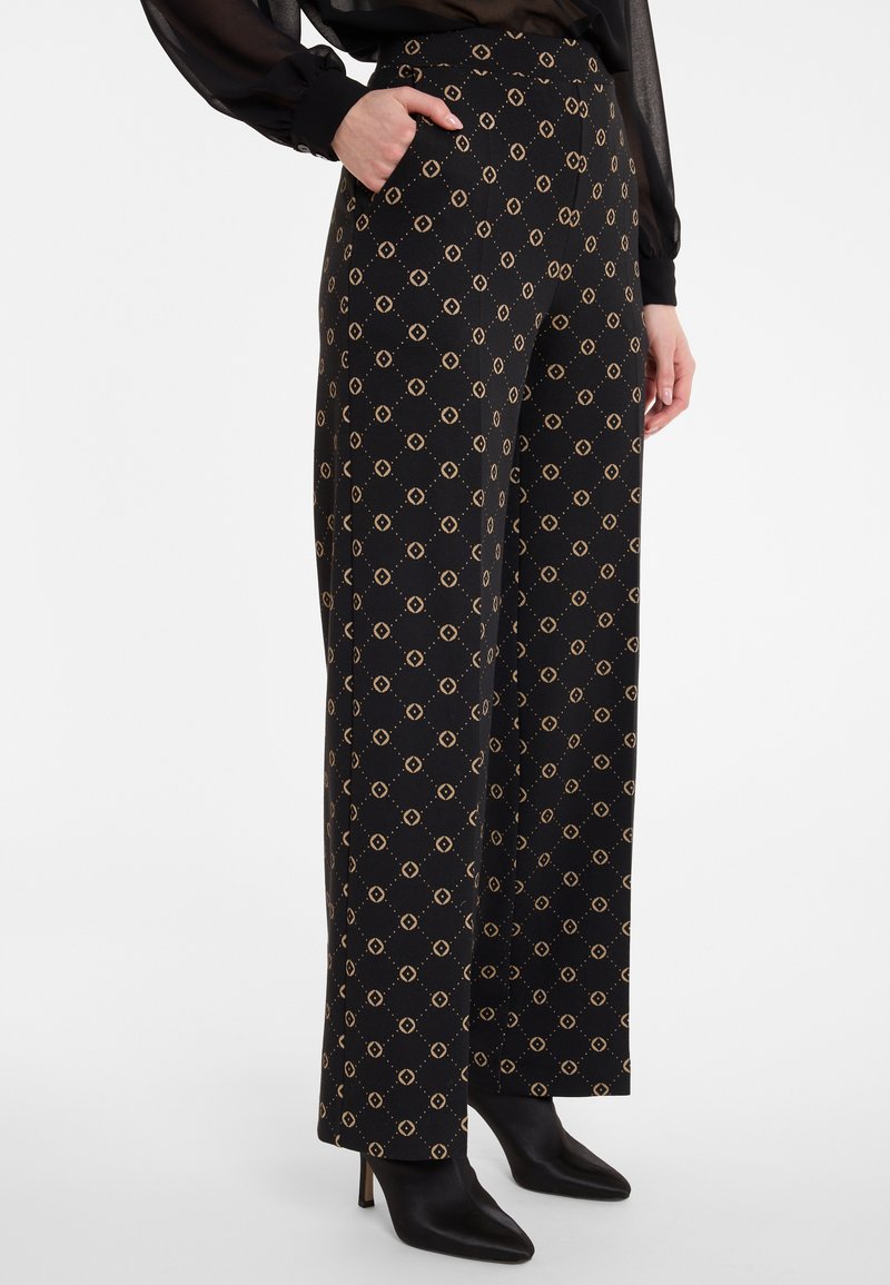 Woman wearing black wide-leg pants with beige geometric pattern, black sheer long-sleeve top, and black heeled ankle boots.