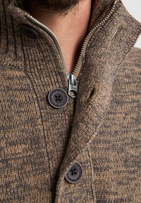 Zbliżenie na dzianinową brązową kurtkę z metalowym zamkiem błyskawicznym i dwoma dużymi czarnymi guzikami w pobliżu kołnierza na szyi osoby.