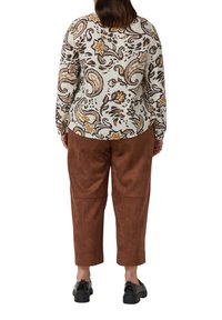 Kobieta z włosami do ramion w kolorze brązowym nosi długą białą bluzkę z brązowym wzorem kwiatowym w stylu paisley, brązowe spodnie i czarne buty, odwrócona plecami.