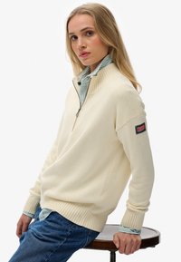 Crème-kleurige gebreide pullover met een hoge kraag en een halve rits, voorzien van geribde manchetten. Een blauw-wit gestreept shirt is eronder gedragen.