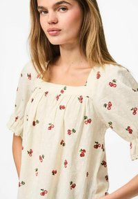 Blusa di crema chiara con scollo quadrato, maniche a sbuffo corte e stampa di ciliegie rosse. Il tessuto testurizzato presenta piccoli punti in rilievo.