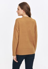 Brązowy sweter z dzianiny z prążkowanym wykończeniem u dołu i na rękawach, o luźnym kroju i prostym okrągłym dekolcie. Noszony z ciemnymi dżinsami.