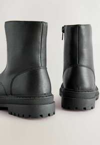 Schwarze Knöchelstiefel aus glattem synthetischem Material, mit einer strukturierten Sohle, runder Zehenpartie und einem seitlichen Reißverschluss zum Schließen.