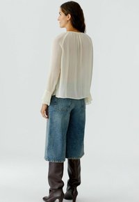 Durchsichtige, cremfarbene Bluse mit langen Ärmeln und Rüschensäumen, kombiniert mit hochgeschnittenen blauen Denim-Culottes und hohen braunen Stiefeln. Einfaches Design, leichte Textur.