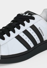 Λευκό αθλητικό παπούτσι Adidas Superstar με μαύρες ρίγες, μαύρα κορδόνια και μαύρη σόλα, με το λογότυπο Adidas στη γλώσσα και το κείμενο «SUPERSTAR» στο πλάι.