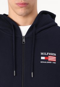 Haut de hoodie zippé marine en tissu doux, avec un logo brodé avec des accents rouges, blancs et bleus. Comprend des cordons de serrage et une fermeture éclair en métal.