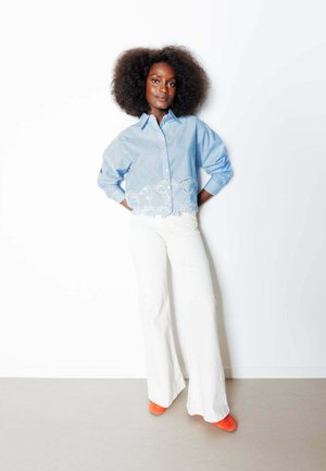 Femme avec une coiffure afro naturelle, debout contre un mur blanc, portant une chemise bleue à rayures avec broderie, un pantalon blanc et des chaussures orange.