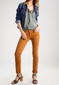 Denim jacket, green shirt, and orange skinny jeans with rolled cuffs. Oblique neckline and heeled sandals with a strap design.