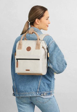 Beige Rucksack mit braunen Lederakzenten, rechteckiger Form, vorderer Reißverschlusstasche und Logo-Patch oben. Der Stoff wirkt robust und strukturiert.