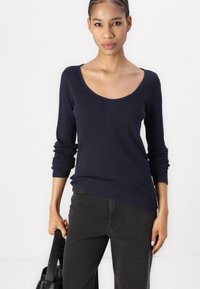Navy ribbed knit top dengan leher scoop dan lengan panjang, dipadukan dengan celana denim gelap. Model memegang tas tangan hitam.