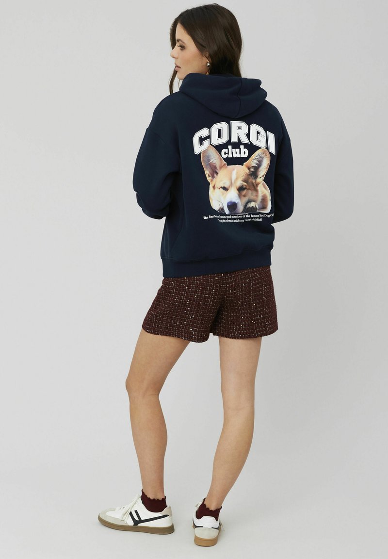 Hoodie marine avec un graphique de Corgi au dos, associé à un short bordeaux texturé et des baskets blanches avec des accents foncés.