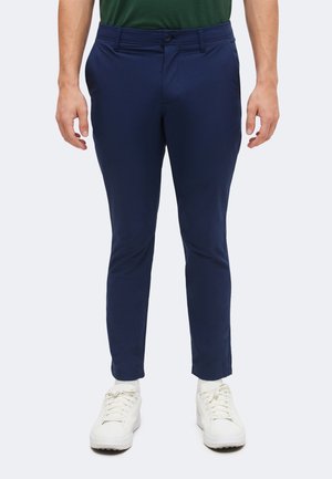 Mand iført navyblå slim-fit bukser med hvide sneakers og en mørkegrøn skjorte, stående mod en ensfarvet lys baggrund.