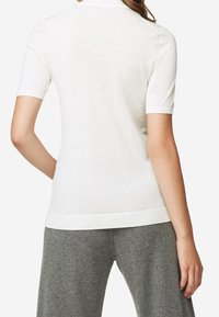 White short-sleeve knitted top with a ribbed neckline and hem. Smooth texture, fitted design, shown worn with grey wool trousers.