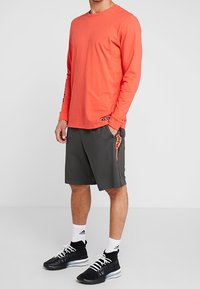 Koszula w kolorze koralowym z długim rękawem, lekkie szare szorty z bocznymi kieszeniami, czarne buty sportowe i białe skarpetki sięgające kostek. Czarne akcenty logo na szortach.