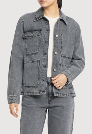 Femme portant une veste en jean noire délavée avec quatre poches avant et un jean assorti, en train de fermer la veste sur un haut blanc.
