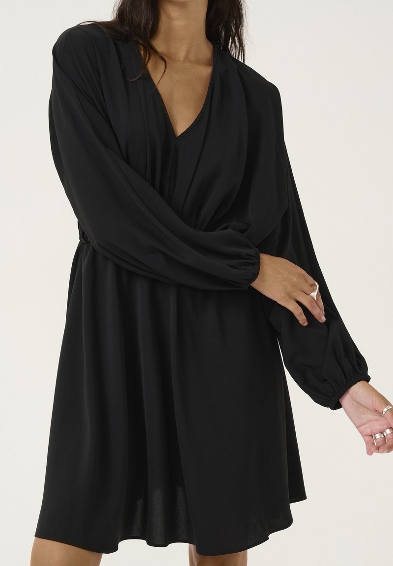 Robe noire avec un profond décolleté en V, manches longues bouffantes et coupe décontractée. Réalisée dans un tissu lisse, elle présente des fronces à la taille.