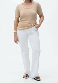 Mujer vestida con un top de manga corta acanalado beige, pantalones de pierna ancha blancos, sandalias negras y una pulsera dorada, de pie con una mano en el bolsillo.