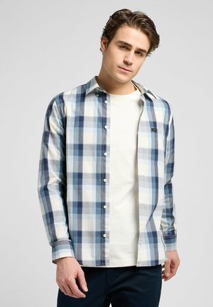 Camisa de cuadros azul y blanca con mangas largas, cuello abotonado y un bolsillo de parche, combinada con una camiseta de color crema debajo.