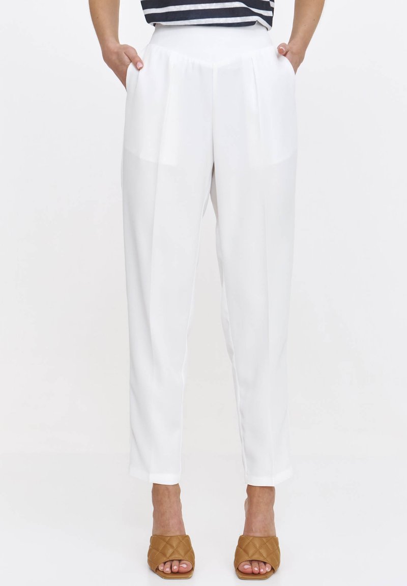 Białe spodniowe spodnie z plisami, bocznymi kieszeniami i gładką fakturą. Stylizowane z brązowymi odkrytymi klapkami, na neutralnym tle.