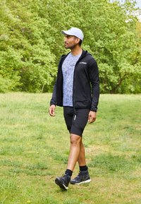 Schwarze Zip-Hoodie über einem hellgrauen gemusterten T-Shirt, kombiniert mit schwarzen Shorts. Schwarze Sportschuhe, die auf Gras in einer grünen Umgebung gehen.