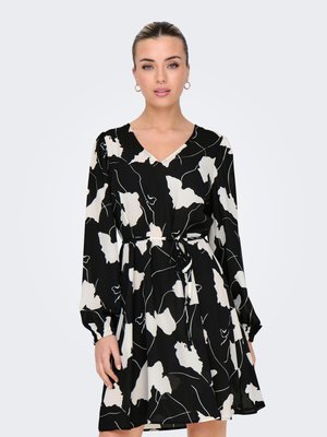 Femme portant une robe à manches longues imprimée de fleurs noire et blanche, avec un décolleté en V et une ceinture à la taille, debout devant un fond clair uni.