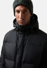 Sort dunjakke med høj krave, trykknaplukning og quiltet design. Kombineret med en mørkegrøn ribstrikket hue med logo.