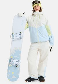Snowboard with white top, blue accents, and splash pattern; person in light blue and white jacket, white pants, and black boots, holding the board.