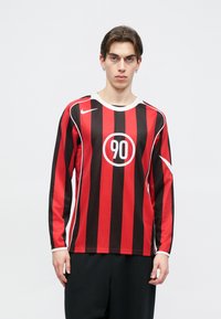 Μακρυμάνικη φανέλα ποδοσφαίρου με κόκκινες και μαύρες κατακόρυφες ρίγες, με στρογγυλή λευκή ετικέτα που έχει τον αριθμό '90' και λευκές λεπτομέρειες.