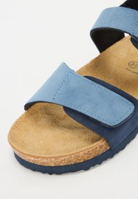 Blå mockaskinnssandaler med två justerbara remmar, korkmellansula och texturerad brun innersula. Har en rundad tå och texturerad yttersula.