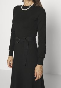 Femme portant une robe noire à manches longues avec une ceinture assortie et un collier de perles, debout devant un fond blanc uni.