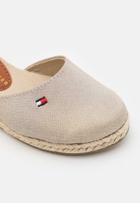 Beżowe espadryle z teksturowanej tkaniny, zamknięty czubek i pleciona podeszwa z juty. Małe haftowane logo w kolorach czerwonym, białym i niebieskim.