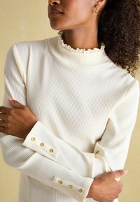 Ribaga kreemjas turtleneck top, millel on frillide äärtega krae ja nööpidega mansetid. Kangas on pehme tekstuuriga, millel on vormistatud disain.