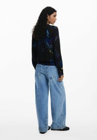 Un pull noir avec des motifs abstraits colorés, associé à un jean en denim bleu clair, ample. Le tissu semble doux et texturé, avec des manches longues.