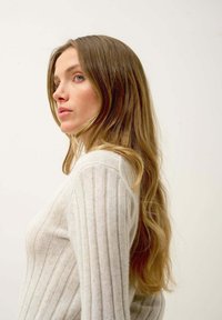 Pull en rib dans un beige clair à manches longues, offrant une texture douce. Le mannequin a de longs cheveux ondulés et est posé de profil sur un fond uni.