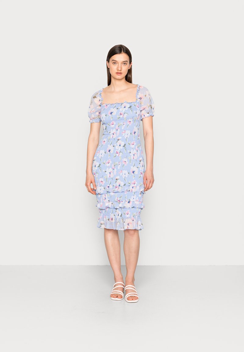 Vestido com padrão floral em azul claro, com mangas bufantes transparentes, corpete ajustado e bainha em camadas com babados. Usado com sandálias brancas com tiras.