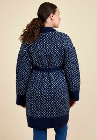 Femeie cu păr creț prins în coadă de cal, purtând un cardigan tricotat bleu închis cu modele albe în formă de diamant și blugi, întorcându-se.