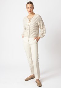 Bež pletivi kardigan s teksturiranim, mjehurastim uzorkom, zatvaranjem na dugmad, u kombinaciji s krem hlačama i taupe slip-on cipelama.