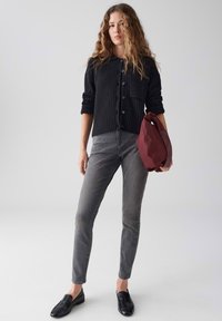 Donna in cardigan nero con bottoni, jeans grigi, mocassini neri, che tiene una borsa piegata bordeaux contro uno sfondo chiaro e uniforme.