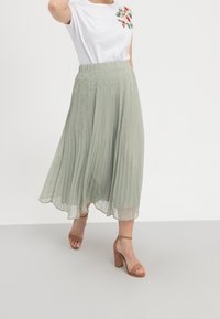 Maxi sukně s plisováním v světle zelené barvě, s plynulým střihem a elastickým pasem. Kombinováno s bílým tričkem s květinovou výšivkou.