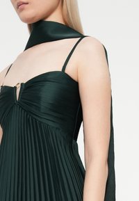Σκούρο πράσινο πλισέ φόρεμα με εφαρμοστό κορσέ, λεπτές τιράντες και ένα αντίστοιχο σατέν μαντήλι κρεμασμένο γύρω από το λαιμό. Εντυπωσιακά χρυσαφένια αξεσουάρ.