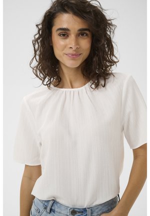 Femme aux cheveux bouclés et foncés portant un chemisier blanc texturé à manches courtes et un jean bleu clair, souriant légèrement sur un fond uni.