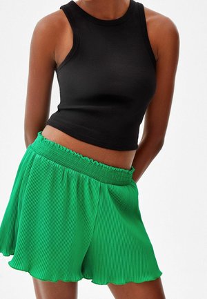 Persona che indossa un top corto nero senza maniche e pantaloncini plissettati verde acceso a vita alta con una fascia arricciata in vita.