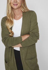 Olive green cardigan with a soft texture, open front, long sleeves, and two front pockets over a white shirt and black bottom.