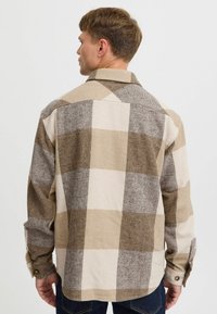 Camicia a quadri marrone e beige realizzata in tessuto morbido, dotata di colletto classico e polsini abbottonati, con un orlo leggermente curvato.